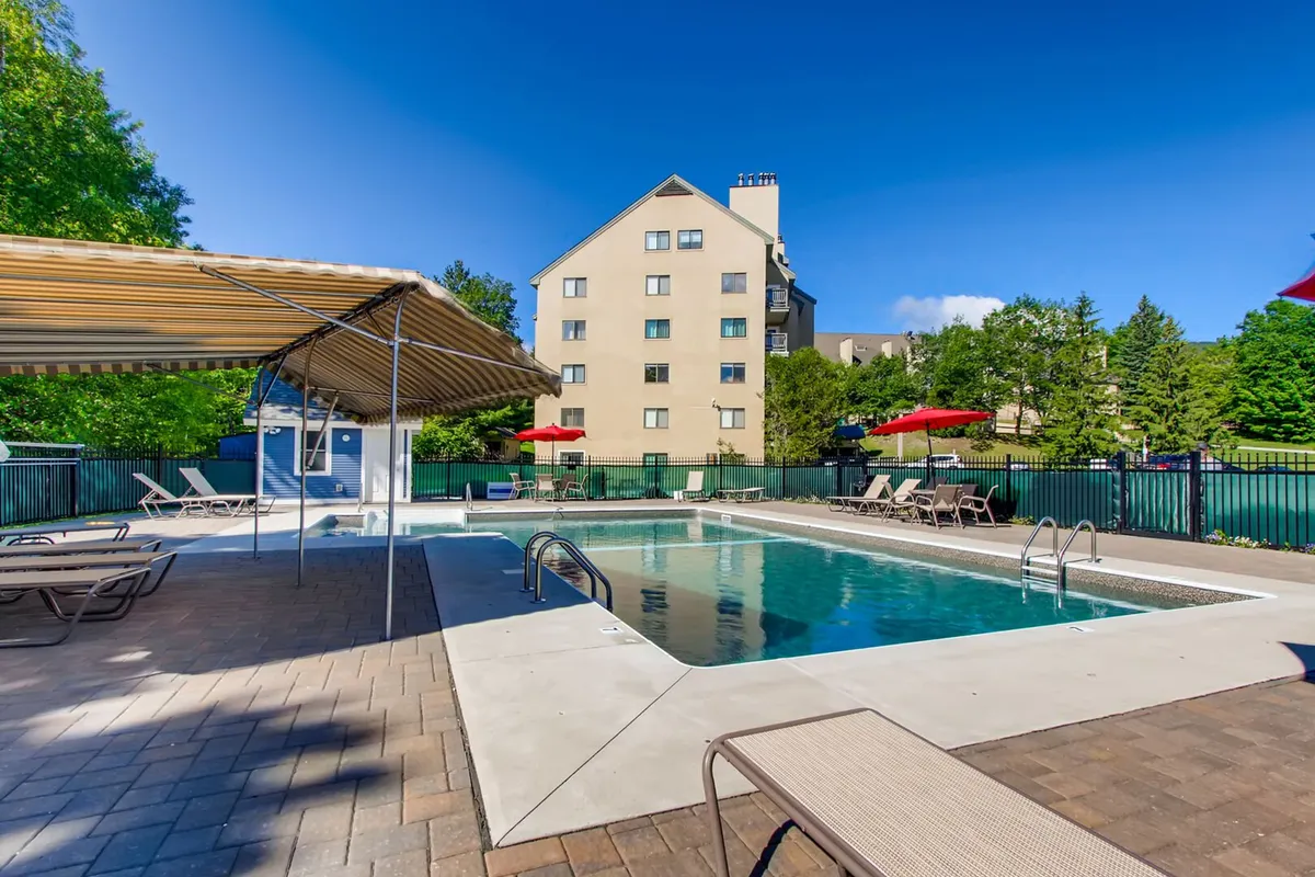 An inviting outdoor swimming pool surrounded by lounge chairs, umbrellas, lush greenery, and a building in the background. The scene is bright and serene, capturing a classic summer resort atmosphere.