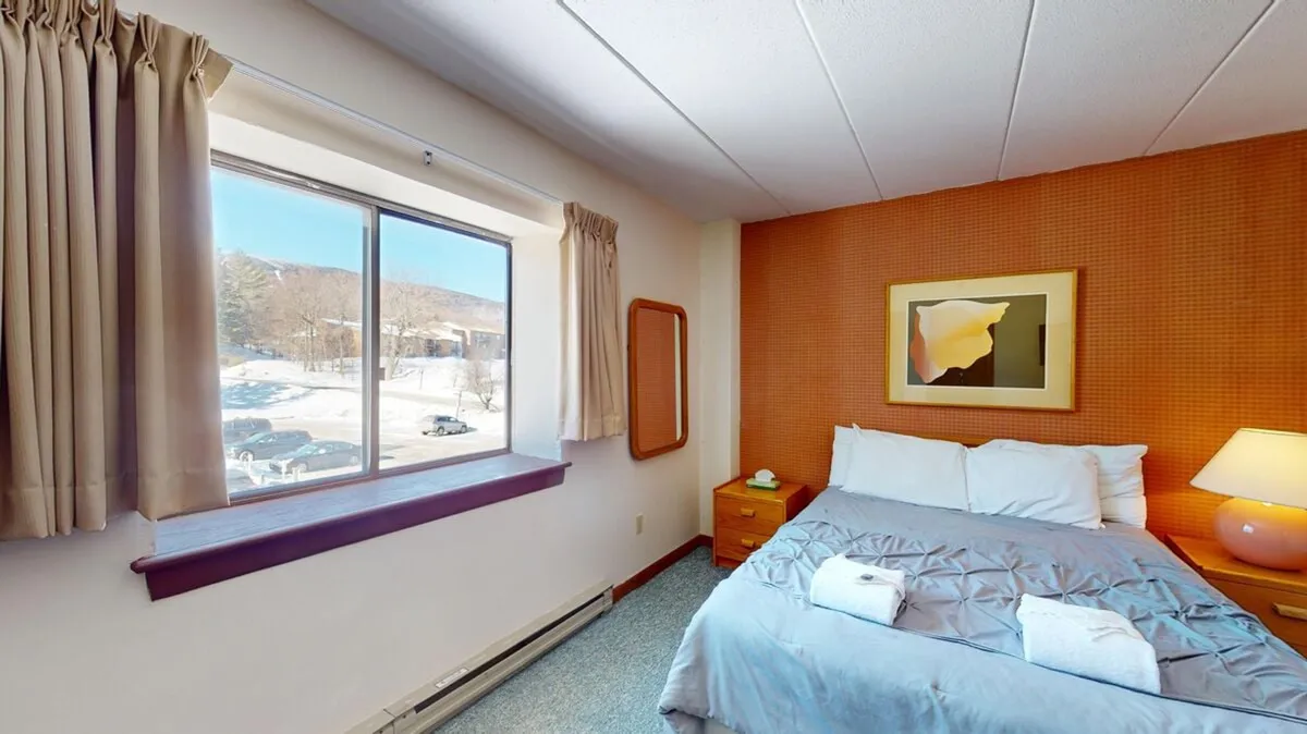 A serene bedroom scene featuring a plush bed dressed in quality linens, a large window letting in natural light, a nightstand with a lamp, and a striking art piece on the wall. There is a cozy atmosphere ideal for relaxing, reading, or enjoying peaceful views.