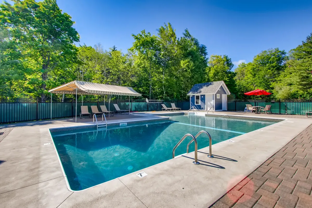 The image shows a vibrant outdoor swimming pool surrounded by lush trees, lounge chairs, a pool house, and a red umbrella. The scene exudes a cheerful, relaxed summer vibe, with space for swimming, sunbathing, relaxing under shade, and socializing.