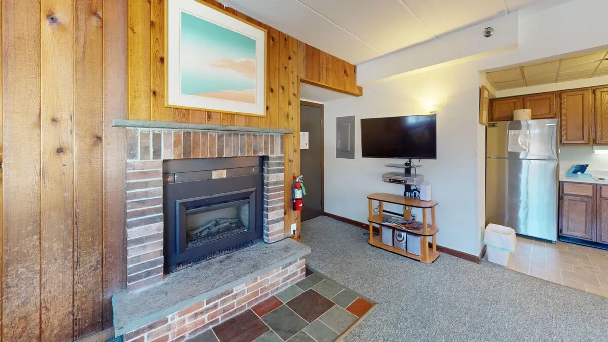 A wide-angle shot of an open-concept living space featuring a living area with a sofa, a wall-mounted television, knotty pine walls, and an adjacent kitchen. Large corner windows allow natural light to fill the space, creating a welcoming, communal environment. The fireplace appears as a centerpiece, even though it’s not currently in use.
