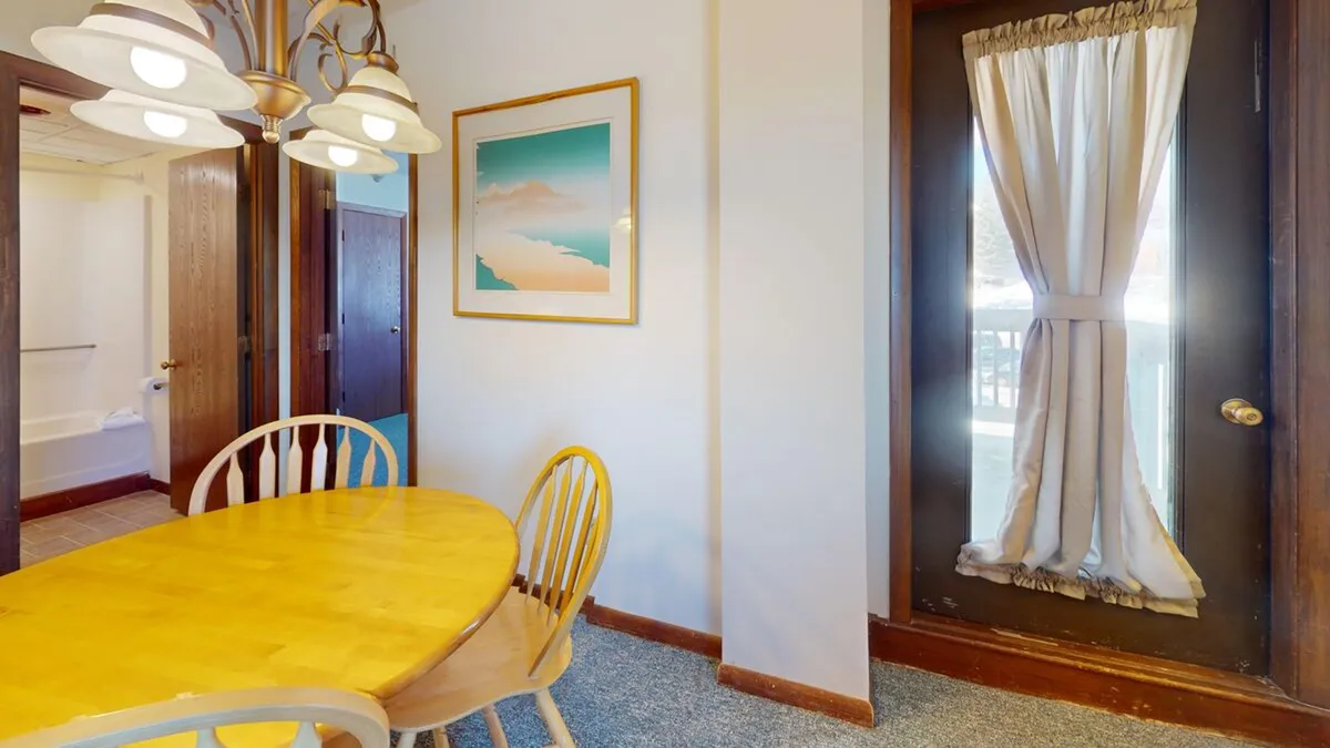 A warmly lit dining area with a central dining table, elegant chandelier, framed artwork, and a doorway with curtains letting in natural light. The space exudes intimacy and comfort, perfect for both breakfasts and evening dinners.