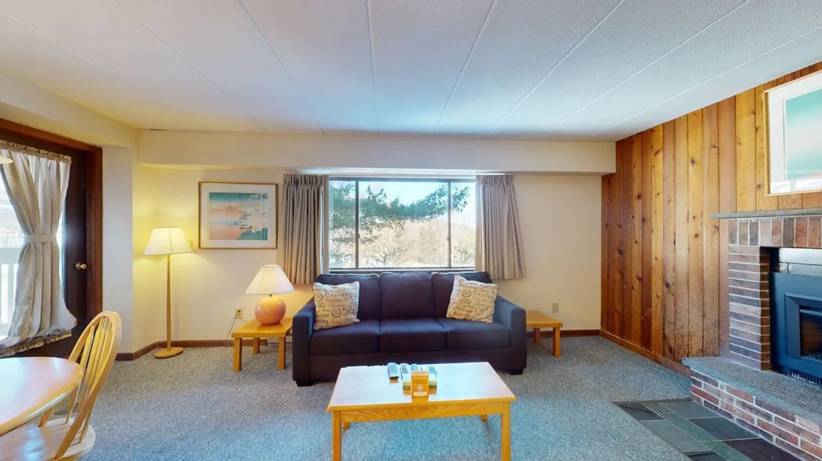 This image displays a cozy living room with a plush sofa, a coffee table, knotty pine walls, a fireplace (currently unavailable, but still present in the image), and a large window letting in ample natural light. The room is tastefully decorated with cheerful, homey accents, and offers a picturesque view through the window.