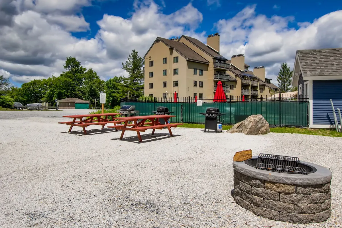 The image highlights an outdoor communal area with picnic tables, grill stations, and a fire pit. A building is visible in the background, indicating proximity to accommodations. The space is designed for barbecuing, picnicking, socializing, and relaxing outdoors—ideal for group gatherings and summer evenings.