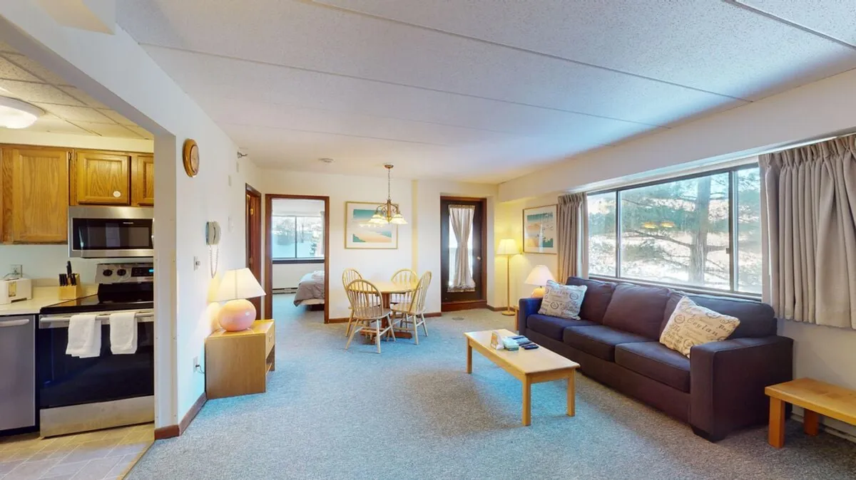 The image showcases a cozy open-concept living space featuring a comfortable sofa, a dining table set for four, a compact but well-equipped kitchen, and large windows that let in natural light. The inviting arrangement suggests an ideal spot for relaxing after a day on the slopes or gathering for a shared meal, with glimpses of the outdoors visible through the window.