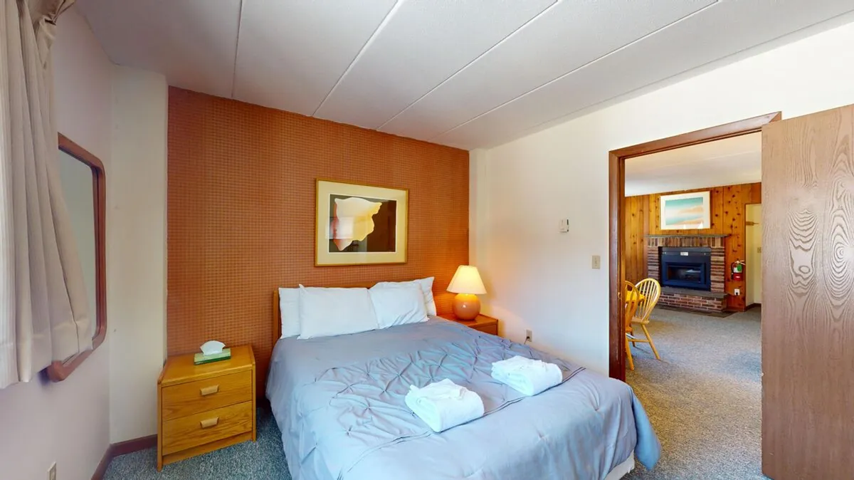 A contemporary bedroom with a comfortable bed, nightstand, lamp, and distinctive wall artwork. The space includes a doorway leading to the living area, suggesting openness and easy flow between rooms. The bedside lamp creates a soft, calming ambiance ideal for nighttime relaxation or quiet reading.